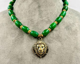May include: A necklace featuring a series of smooth, oval-shaped green beads separated by small gold-toned spacers. The centerpiece is a circular gold-toned pendant depicting a detailed lion head in relief, hanging from the green bead strand.