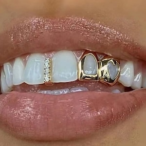 Puede incluir: Un conjunto de grill dental de oro que presenta una barra vertical con pequeñas piedras transparentes en un diente y una funda de oro con un recorte en forma de corazón en el diente adyacente, sobre los dientes superiores de esmalte blanco.