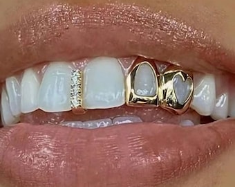 May include: A gold tooth grill set featuring a vertical bar with small clear stones on one tooth and a gold cap with a heart-shaped cutout on the adjacent tooth. The dental jewelry is positioned on the upper teeth, contrasting with the natural white enamel.