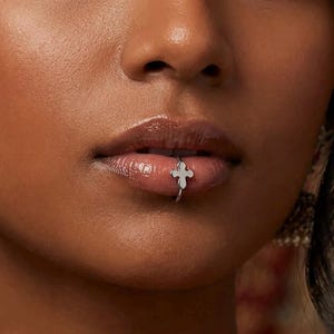 May include: A silver-toned lip ring featuring a small, four-pointed cross charm. The jewelry is positioned on the lower lip, with the thin metal band encircling the lip tissue. The cross charm rests against the outer surface of the lower lip.