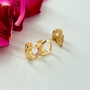 May include: Two gold-toned tooth grills. One piece features a heart-shaped cutout design, while the other is a single tooth cap with a textured, grid-like surface and a vertical row of small, clear, sparkling stones set along the side edge.
