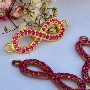 May include: Gold-toned infinity charm with a vibrant pink gemstone setting. The charm features a classic infinity symbol design, with a loop on each end for attachment. The gemstones are small and closely set, creating a continuous line of color.