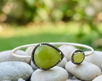 May include: A silver open cuff bracelet featuring a large pear-shaped faceted lime green stone and a smaller round faceted lime green stone, both set in dark metal frames with small dark stone accents along the edges.