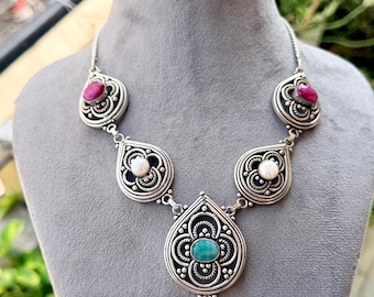 May include: A silver necklace featuring five teardrop-shaped pendants with intricate metalwork. The central pendant holds a green stone, two side pendants contain white pearls, and the two outer pendants feature small, vibrant pink stones.