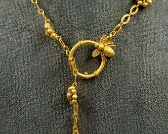 May include: A gold-toned necklace featuring a central circular toggle clasp adorned with a small bee figure. The chain is embellished with spaced floral-shaped clusters of tiny spheres, creating a delicate and intricate design throughout the piece.