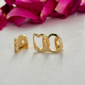 May include: A set of gold-toned tooth grills featuring a polished finish. One piece displays a heart-shaped cutout, while the other piece is accented with a vertical row of small, clear, sparkling stones along the side edge of the metal frame.