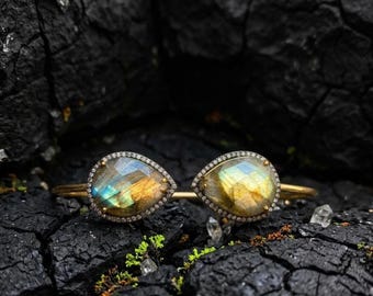May include: A gold cuff bracelet featuring two pear-shaped labradorite gemstones, each surrounded by a halo of small white pavé diamonds. The labradorite stones exhibit iridescent flashes of blue and yellow, set in a polished gold metal band.