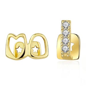 May include: Two gold-tone tooth grills. The left piece features two open heart-shaped frames with star-shaped cutouts. The right piece is a vertical bar set with four clear round stones, attached to a square base with a star-shaped cutout.