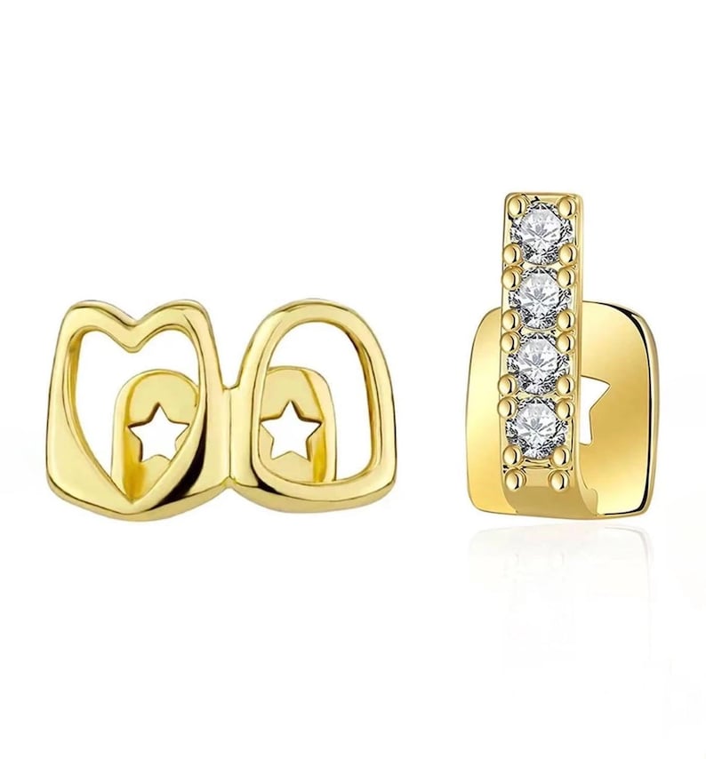 May include: Two gold-tone tooth grills. The left piece features two open heart-shaped frames with star-shaped cutouts. The right piece is a vertical bar set with four clear round stones, attached to a square base with a star-shaped cutout.