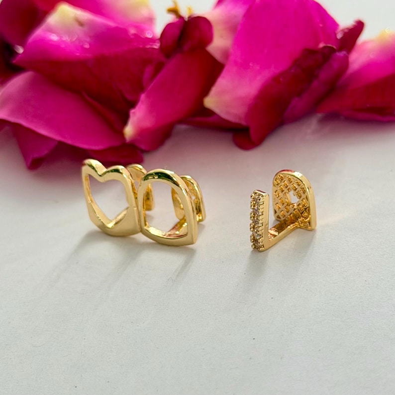 May include: Two gold-toned tooth grills. One piece features a hollow heart-shaped design, while the other is a single tooth cap with a vertical row of small clear stones and a textured grid pattern on the side surface, set against pink petals.