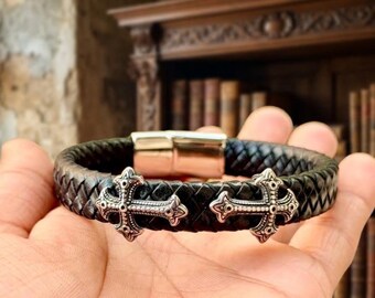 May include: A black braided leather bracelet featuring two silver-toned metal cross charms and a polished rectangular magnetic clasp. The bracelet has a textured woven surface and is designed with a symmetrical arrangement of the cross ornaments.