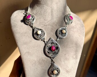 May include: A silver necklace featuring a central teardrop pendant with a red stone and a smaller dangling pearl pendant. The chain includes additional teardrop-shaped links decorated with intricate metalwork, two red stones, and two white pearls.