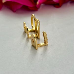 May include: Two gold-toned ear cuffs with a polished finish. The front ear cuff features a small star cutout on the side and a row of tiny clear stones along the edge. The second ear cuff is a simple, smooth, open-ended band design.