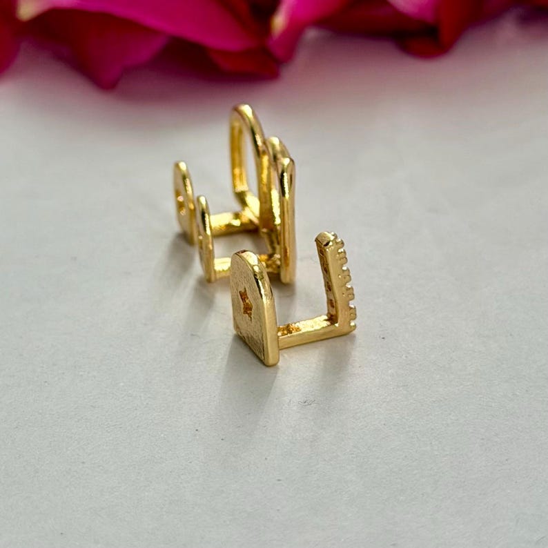May include: Two gold-toned ear cuffs with a polished finish. The front ear cuff features a small star cutout on the side and a row of tiny clear stones along the edge. The second ear cuff is a simple, smooth, open-ended band design.