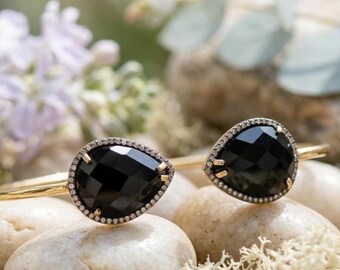 May include: A gold cuff bracelet featuring two teardrop-shaped black faceted stones, each surrounded by a delicate halo of small white pavé diamonds. The open-ended design showcases a polished gold band that connects to the stone settings.
