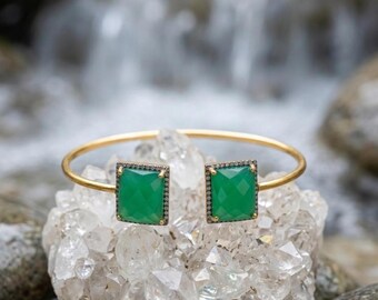 May include: A gold-toned open cuff bracelet featuring two rectangular, faceted green stones. Each stone is framed by a border of small, dark, pavé-set crystals. The bracelet has a thin, smooth, circular band that connects to the stone settings.