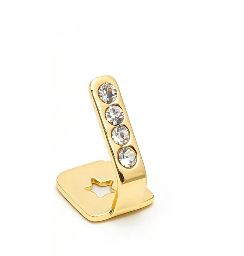 May include: A gold-toned metal accessory featuring a flat square base with a star-shaped cutout and an angled upright arm. The upright arm is embellished with four clear, round, faceted crystals arranged in a vertical line along its surface.