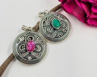 May include: Two circular silver pendants featuring intricate filigree scrollwork and beaded detailing. One pendant is set with a central oval pink gemstone, while the other features a central oval green gemstone, both with a polished finish.
