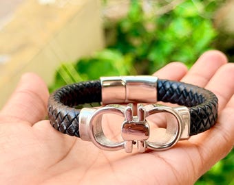 May include: A black braided leather bracelet featuring a polished silver-tone metal clasp with a symmetrical double-loop design. The bracelet has a secure magnetic closure mechanism and a textured woven band that wraps around the wrist.