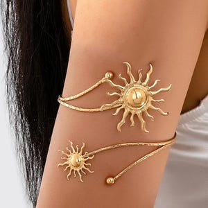 May include: A gold-toned metal arm cuff featuring two sun motifs of different sizes. The design includes a textured, spiraling band that wraps around the arm, ending in small spherical beads at each terminal point of the metal wire structure.