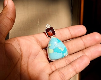 May include: A pendant featuring a triangular turquoise stone with light blue and green veining set in silver, topped with a smaller square faceted dark red garnet stone. The pendant has a silver bail at the top for attachment to a chain or cord.