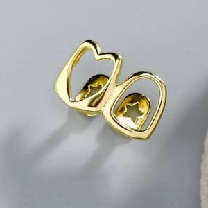 May include: A gold-colored dental grill for two teeth, featuring a polished metallic finish and a cutout star shape centered within each tooth frame. The design consists of two connected, rounded rectangular outlines with open centers.