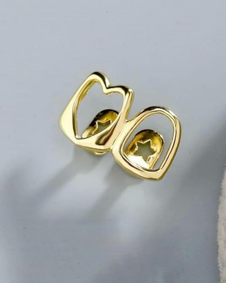 May include: A gold-colored dental grill for two teeth, featuring a polished metallic finish and a cutout star shape centered within each tooth frame. The design consists of two connected, rounded rectangular outlines with open centers.