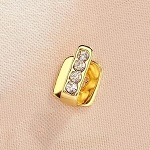 May include: A gold-toned ear cuff featuring a rectangular base and a vertical bar set with four clear, round-cut rhinestones. The piece has a polished metallic finish and a simple open design, showcasing a clean, reflective surface and clear stones.