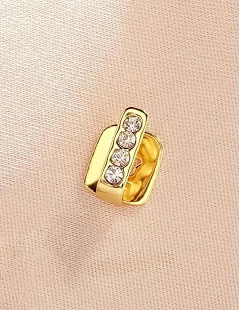 May include: A gold-toned ear cuff featuring a rectangular base and a vertical bar set with four clear, round-cut rhinestones. The piece has a polished metallic finish and a simple open design, showcasing a clean, reflective surface and clear stones.
