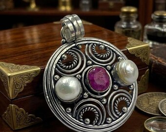 May include: A circular silver pendant featuring an intricate openwork design with twisted wire accents. The center holds a round, faceted deep pink ruby, flanked by two smooth, white, round pearls set within the silver frame and a top bail.