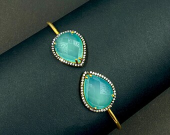 May include: A gold-toned open cuff bracelet featuring two teardrop-shaped, translucent light blue stones. Each stone is framed by a border of small, clear pavé-set crystals, creating a symmetrical design that highlights the smooth, polished finish.