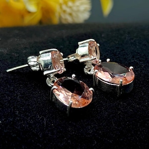 Puede incluir: Pendientes de plata con piedras preciosas ovaladas de color melocotón. Los pendientes tienen un diseño de poste y tachuela, con una piedra preciosa más pequeña en el poste y una piedra preciosa colgante más grande debajo. Fondo negro.