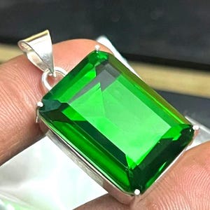 Puede incluir: Un colgante rectangular de piedra preciosa verde esmeralda con un engaste de plata. La piedra preciosa facetada refleja la luz y está suspendida de una fianza de plata. El colgante se sostiene entre los dedos.