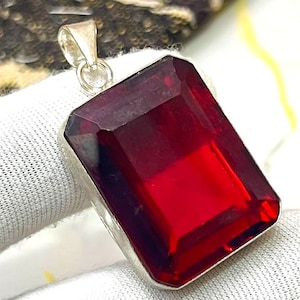 Peut inclure: Pendentif rectangulaire en pierre précieuse rouge foncé avec une bélière argentée. La pierre précieuse est facettée, captant la lumière et affichant une couleur riche et saturée. Le pendentif est probablement un bijou, peut-être un collier.