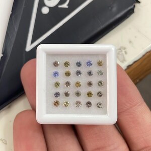 May include: A white plastic tray filled with small, round gemstones in various colors, including yellow, blue, and clear. The tray is held in a hand, with a black box bearing the letters "S.S." in the background.