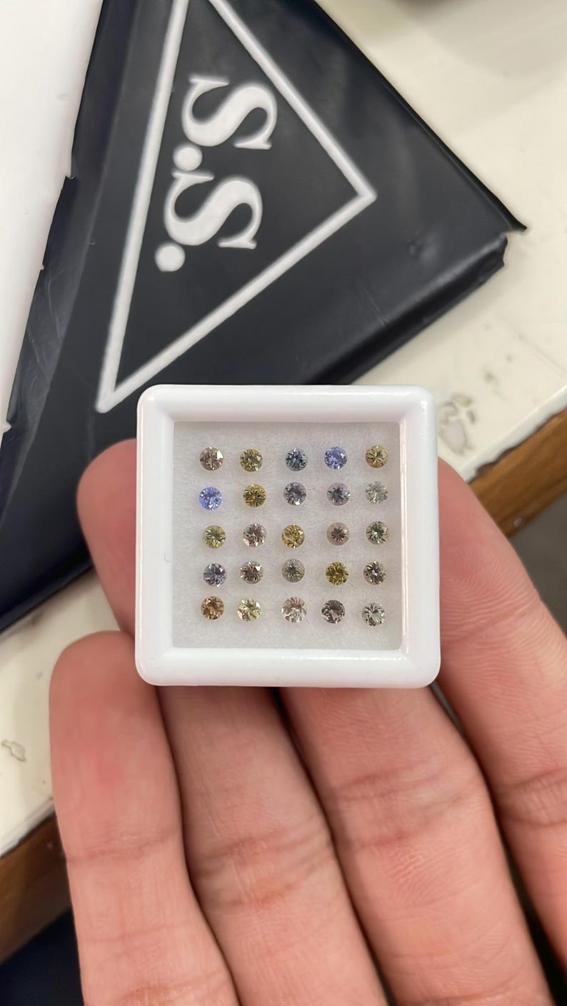 May include: A white plastic tray filled with small, round gemstones in various colors, including yellow, blue, and clear. The tray is held in a hand, with a black box bearing the letters "S.S." in the background.