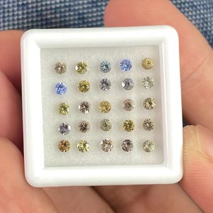 May include: A white square tray filled with twenty-four small, round gemstones in various colors. The gemstones include shades of yellow, blue, brown, and gray. The tray is held in a person's hand.