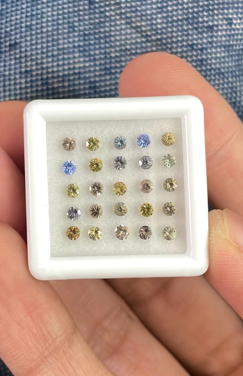 May include: A white square tray filled with twenty-four small, round gemstones in various colors. The gemstones include shades of yellow, blue, brown, and gray. The tray is held in a person's hand.