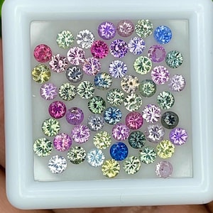 Op de afbeelding: Een verzameling kleine, ronde, gefacetteerde edelstenen in diverse kleuren, waaronder roze, paars, blauw, groen en geel, gerangschikt in een vierkante witte displaydoos met een doorzichtig deksel.