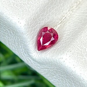 May include: A pear-shaped, faceted ruby gemstone with a deep red color and a brilliant, reflective surface. The stone features a pointed top and a rounded base, showcasing precise geometric cuts that enhance its natural clarity and vibrant hue.