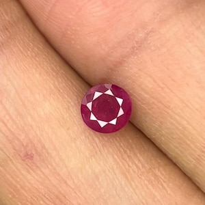 May include: A round, faceted, deep pinkish-red gemstone with a star-like pattern visible in the center. The stone features a brilliant cut with multiple triangular facets radiating from the middle, creating a distinct geometric light reflection.