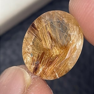 Puede incluir: Una piedra preciosa de cuarzo rutilado facetada de forma ovalada, con un cuerpo claro y transparente lleno de numerosas inclusiones de color marrón dorado en forma de aguja que irradian por el interior de la piedra.