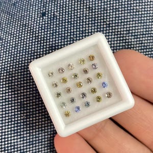 May include: A white square tray filled with small, round gemstones in various colors, including clear, yellow, purple, and blue. The gemstones are set against a white background, and the tray is held in a hand.