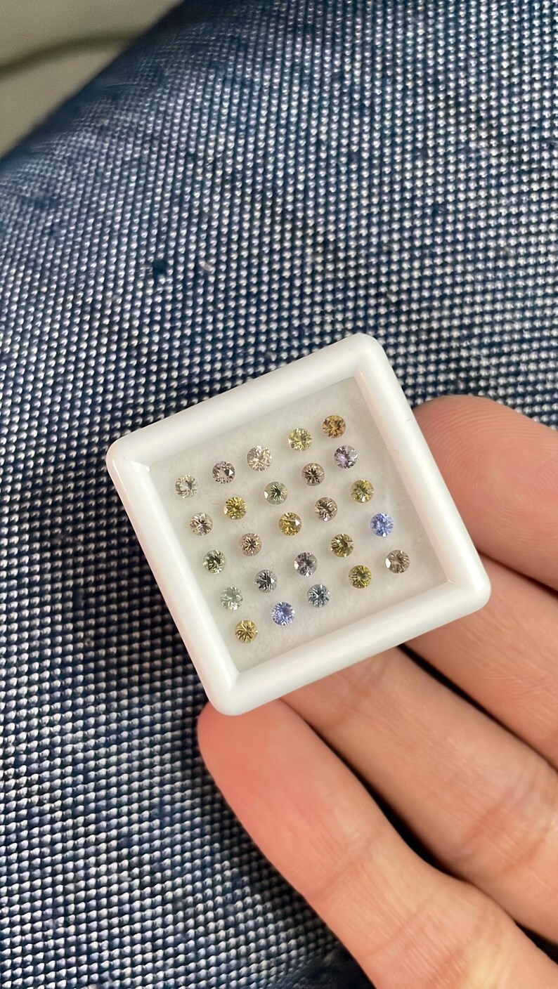 May include: A white square tray filled with small, round gemstones in various colors, including clear, yellow, purple, and blue. The gemstones are set against a white background, and the tray is held in a hand.