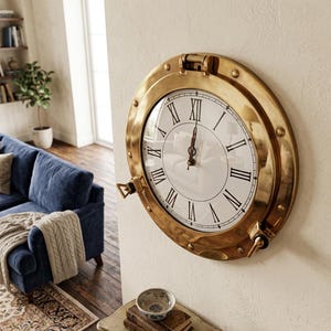 May include: A circular wall clock with a polished brass frame designed like a nautical porthole. The clock face is white with black Roman numerals and two thin black hands. The frame features a hinged top and side locking mechanisms.