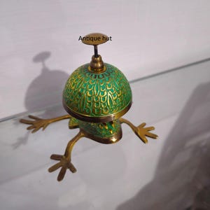 以下が含まれることがあります： 真鍮製のカエル型のベルで、緑と金の模様のドームが付いています。「Antique hut」と書かれた小さなハンドルが上部にあります。カエルの足は伸びており、風変わりな外観になっています。