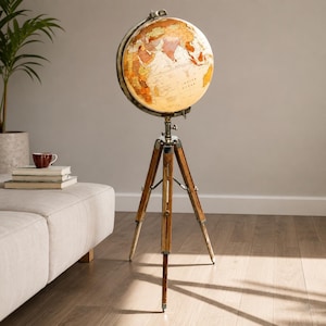 Pode incluir: Um globo terrestre iluminado de chão com um mapa em tons quentes de âmbar, bege e creme. A esfera está montada num anel meridiano metálico e apoiada por uma base de tripé de madeira de três pernas com ferragens prateadas.