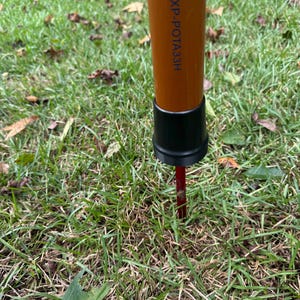 Puede incluir: Un poste naranja con el texto "ZXP-POTA33H" se inserta en el suelo. El poste tiene una base negra y una estaca roja. La imagen está tomada desde un ángulo bajo, mostrando el poste en una zona de hierba.