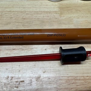 Puede incluir: Una herramienta roja y negra con un extremo puntiagudo, probablemente para estacar o marcar. La herramienta tiene un mango negro y un eje largo y rojo. En el fondo hay un tubo marrón con texto. El texto dice "Portable Telescopic Carbon Fiber Mast" y "ZXP-POTA33".