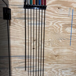May include: A black metal tool holder mounted on a wood surface, holding eight long, thin tools. The tools have black, gray, green, orange, black, blue, and red handles. The tools are evenly spaced and appear to be in a workshop or garage setting.